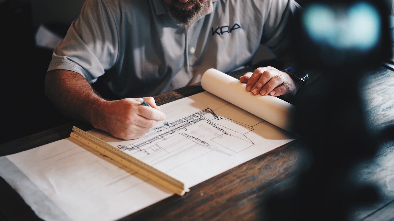 An architect drawing a building elevation by hand, with a scale rule and rolled drawings on the desk